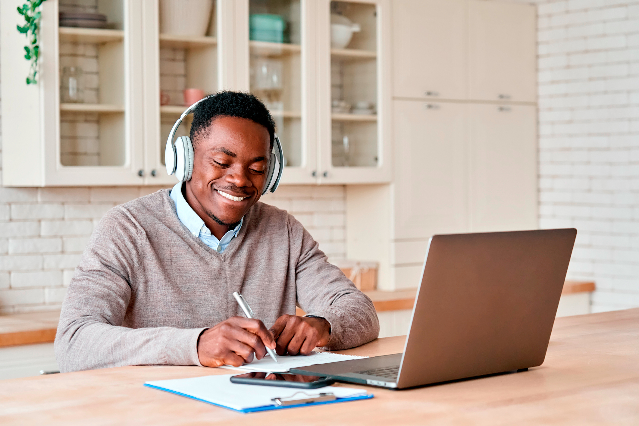 Happy african business man, black male student wearing headphones elearning on laptop computer sitting at kitchen table working from home office, learning online, studying remote training course.