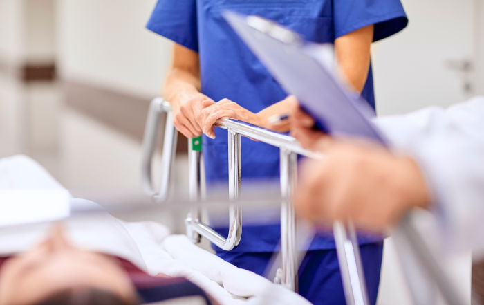 Photo of a person mostly off screen in a white lab coat holding a clipboard next to a hospital bed where a person is lying down